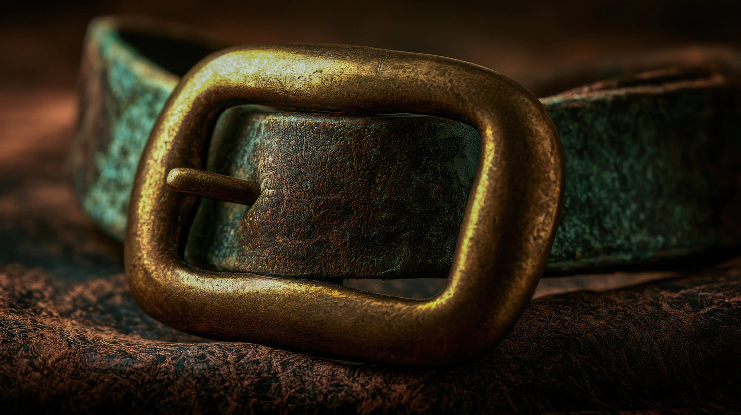 A dramatic, extreme close-up photograph highlighting the rugged hardware of a Caracoda Equestrian heirloom piece. A heavy, solid brass buckle with a warm, worn patina secures a thick strap of heavily weathered vintage saddle leather. The leather exhibits deep textural scarring and striking traces of authentic verdigris, showcasing years of enduring character. Moody, directional lighting emphasises the robust, time-honoured craftsmanship and enduring quality of our traditional English leatherwork.