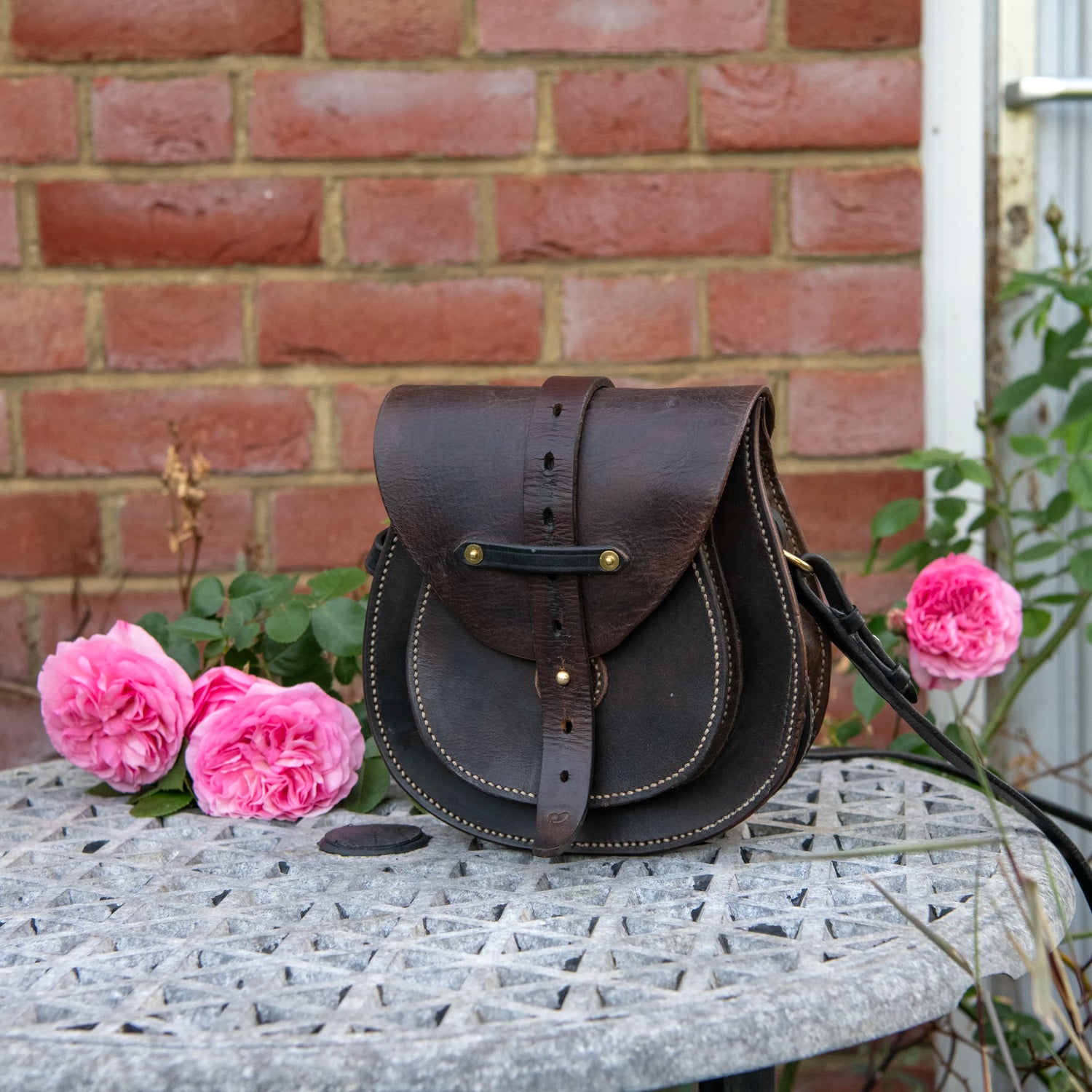 A close-up environmental photograph of The Uffington restored leather scrip resting on a stone garden table against a traditional red brick wall. Framed by blooming pink roses, the tight angle highlights the intricate hand-driven saddle stitching, the natural conditioning of the vintage saddle leather, and the horizontal keeper strap secured by twin solid brass rivets across the main flap.