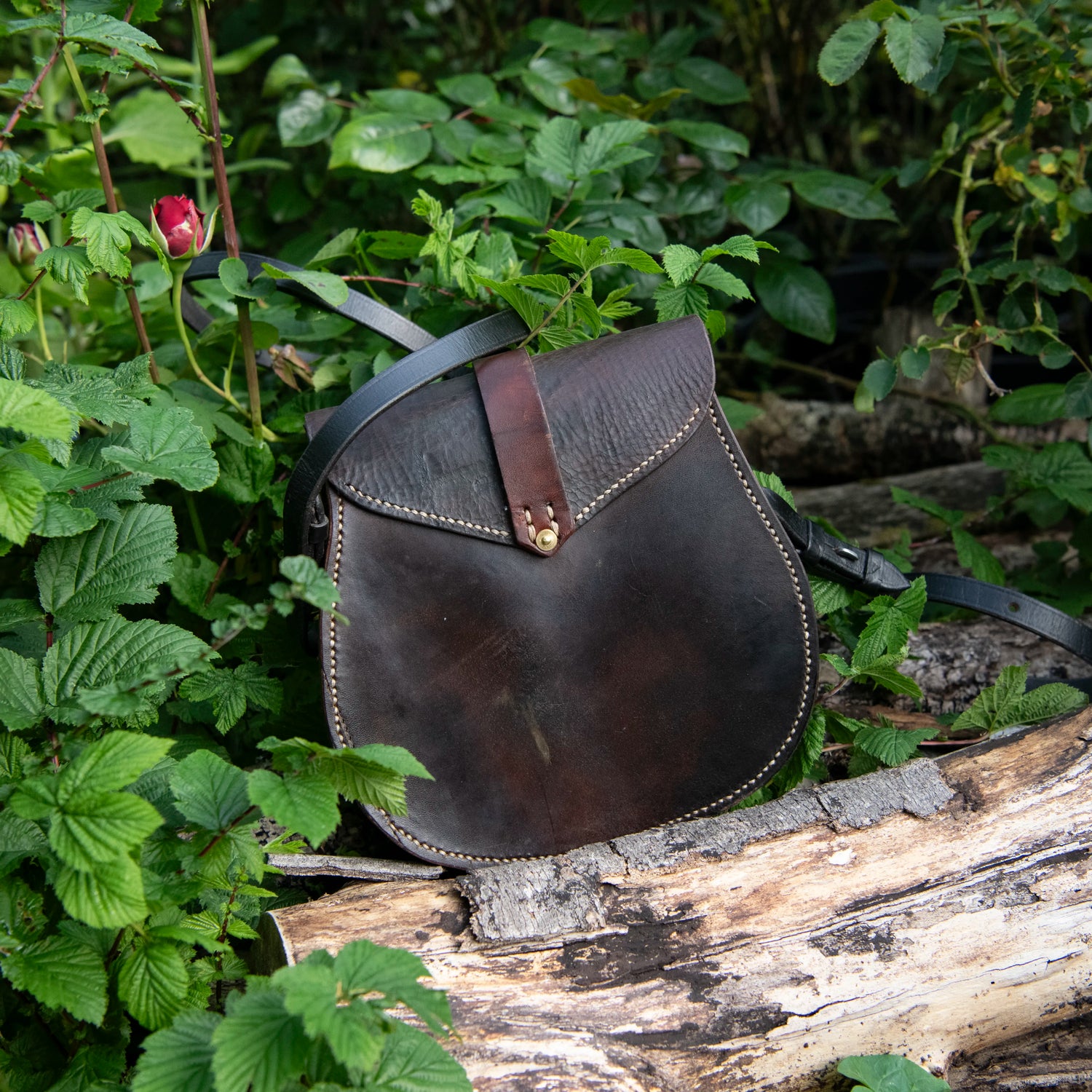 A moody, earthy photograph capturing the rear profile of The Uffington leather scrip resting heavily upon a weathered timber log. Surrounded by green foliage and a subtle red rosebud, the image highlights the unbroken expanse and rich patina of the restored dark saddle leather. The structural horseshoe silhouette is framed by traditional hand-driven saddle stitching and secured with a solid brass stud.