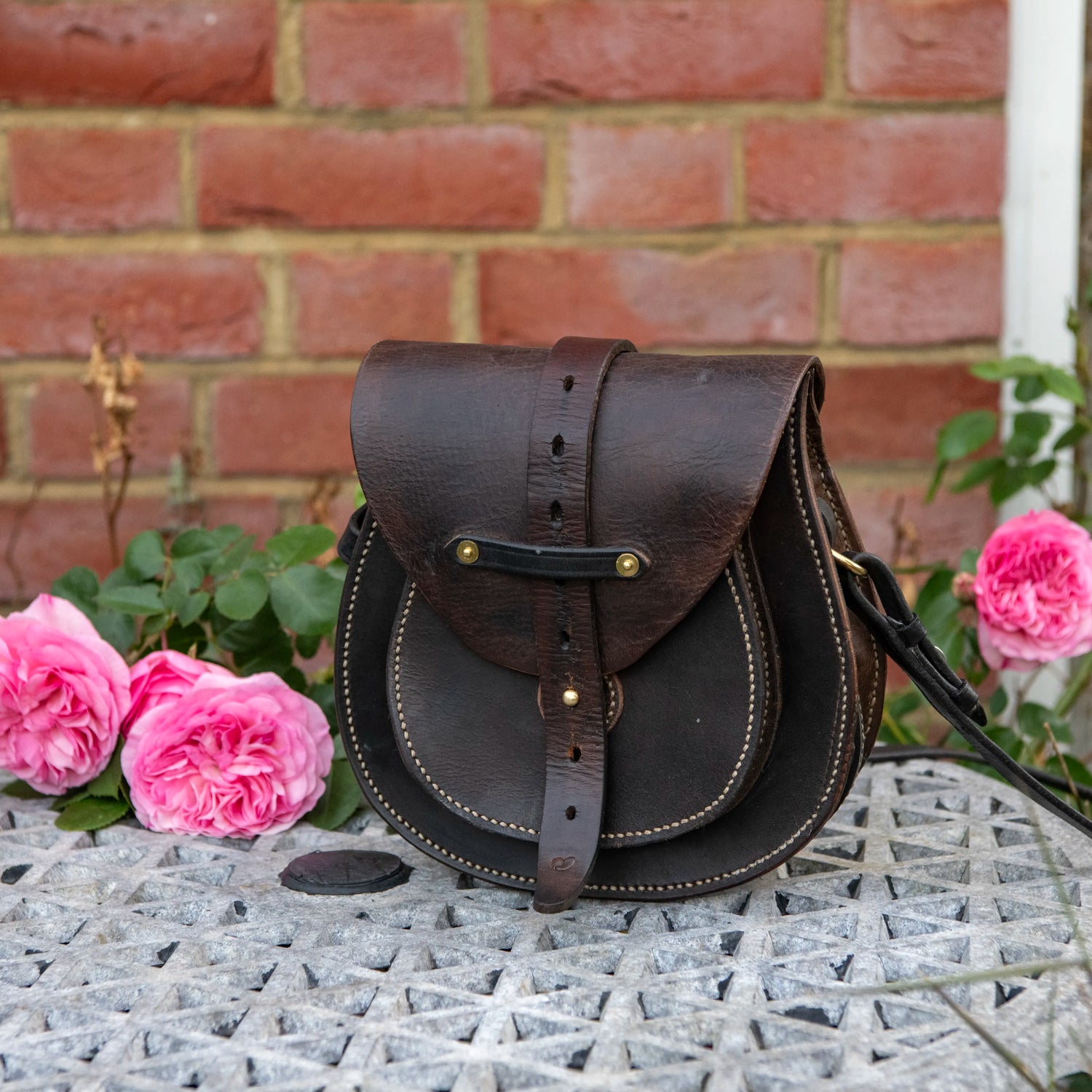 An environmental photograph of The Uffington leather scrip resting upright on a textured stone garden table. Set against a traditional English red brick wall and framed by vibrant pink roses, the dark, restored saddle leather displays immense visual weight. The image clearly shows the layered doublet construction, the scaled-down front pocket, and the heavy central harness strap secured by a brass Sam Browne stud.