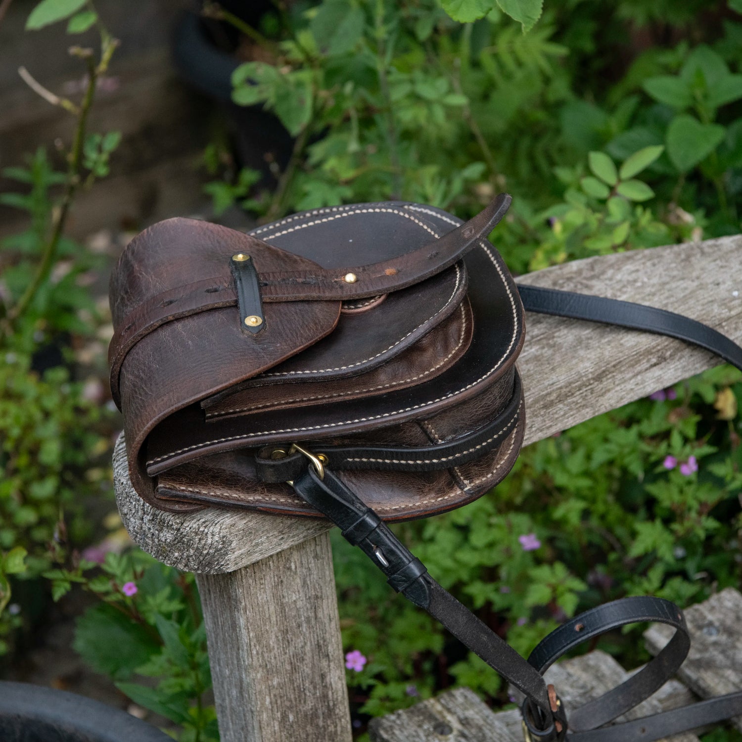 A side-profile environmental photograph of The Uffington restored leather scrip resting on a weathered wooden post. Framed by green foliage, the angle clearly illustrates the physical depth of the side gusset and the layered doublet construction. The thick, dark saddle leather flap curves over the front pocket, while the heavy black equestrian shoulder strap drops down into the foreground, securely anchored by a solid brass D-ring.