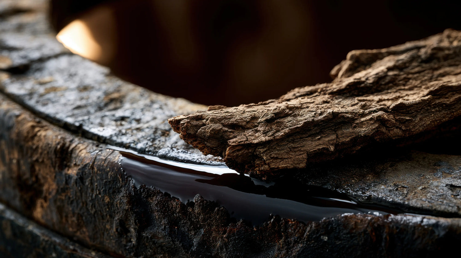 A sharp, close-up macro photograph of raw, deeply textured oak bark resting on the weathered, dark timber edge of a traditional leather-tanning vat at Caracoda Equestrian. A pool of rich, dark tanning liquor clings to the wood, highlighting the authentic, time-honoured process of natural oak bark tanning used to prepare our heritage saddle leather.