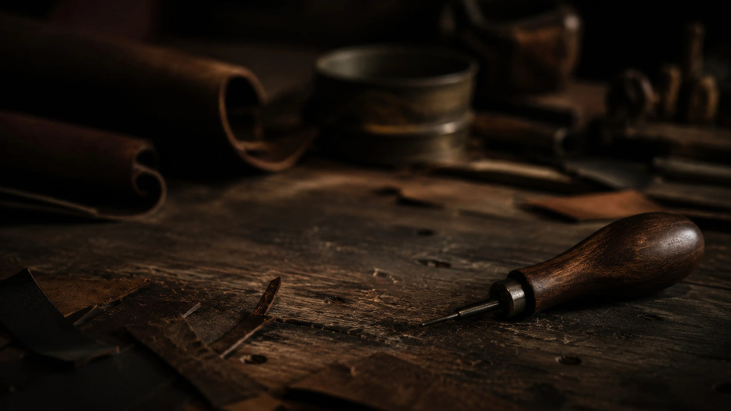 A close-up, moody photograph of a traditional wooden-handled leather-working awl resting on a weathered, rustic timber workbench in the Caracoda Equestrian workshop. Surrounded by raw leather offcuts, the scene captures the meticulous, hand-crafted restoration process of vintage English saddlery.