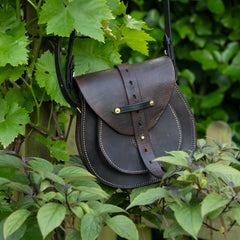A moody, natural light photograph of The Uffington bag, crafted from restored, dark vintage saddle leather, hanging with palpable visual weight amidst lush green vine leaves and foliage. The traditional saddle stitching and brass hardware glint against the deep shadows of the leather.