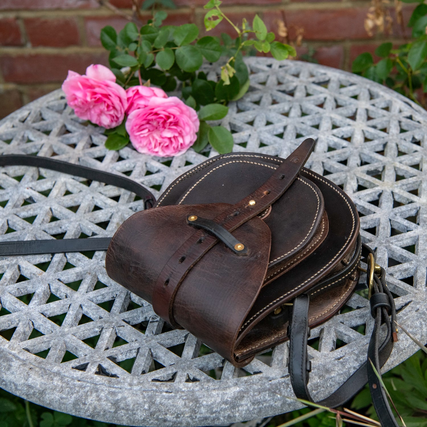A high-angle environmental photograph of The Uffington restored leather scrip lying horizontally on a textured stone garden table. Framed by blooming pink roses and a traditional red brick wall, this reclined perspective perfectly demonstrates the architectural depth of the doublet construction. The heavy, dark saddle leather flap curves over the structured front pocket, highlighting the central harness strap, the natural grain of the vintage salvage, and the solid brass hardware.