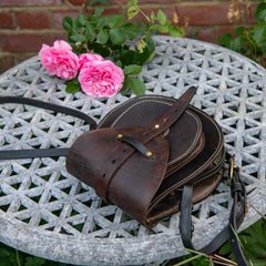 A high-angle environmental photograph of The Uffington restored leather scrip lying horizontally on a textured stone garden table. Framed by blooming pink roses and a traditional red brick wall, this reclined perspective perfectly demonstrates the architectural depth of the doublet construction. The heavy, dark saddle leather flap curves over the structured front pocket, highlighting the central harness strap, the natural grain of the vintage salvage, and the solid brass hardware.