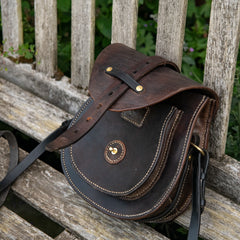 An environmental photograph of The Uffington restored leather scrip resting on a weathered wooden garden bench. The heavy saddle leather main flap is lifted, revealing the stitched Caracoda maker's mark on the inner panel. The image focuses on the layered front pocket, the hand-driven saddle stitching, and the solid brass Sam Browne stud used to secure the bag, all set against a backdrop of rustic timber slats and green foliage.