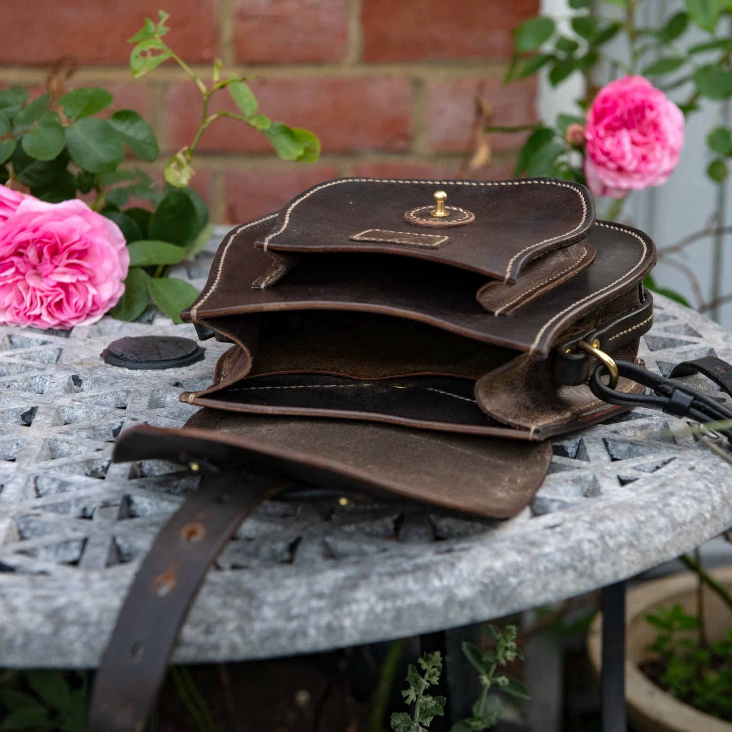 An environmental photograph showing The Uffington restored leather scrip laid fully open on a stone garden table against a red brick wall with pink roses. The main heavy saddle leather flap is drawn back, revealing the unlined, natural interior of the main compartment and the layered doublet construction. The structured front pocket is prominently displayed, secured by a central solid brass Sam Browne stud.