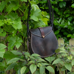 A moody, natural light photograph showing the rear profile of The Uffington leather scrip. Hanging amidst vibrant green foliage, the broad back panel of the bag displays the rich, glass-like patina of restored vintage saddlery. The heavy visual weight of the dark equestrian leather is accentuated by the meticulous hand-driven saddle stitching and the robust central strap.
