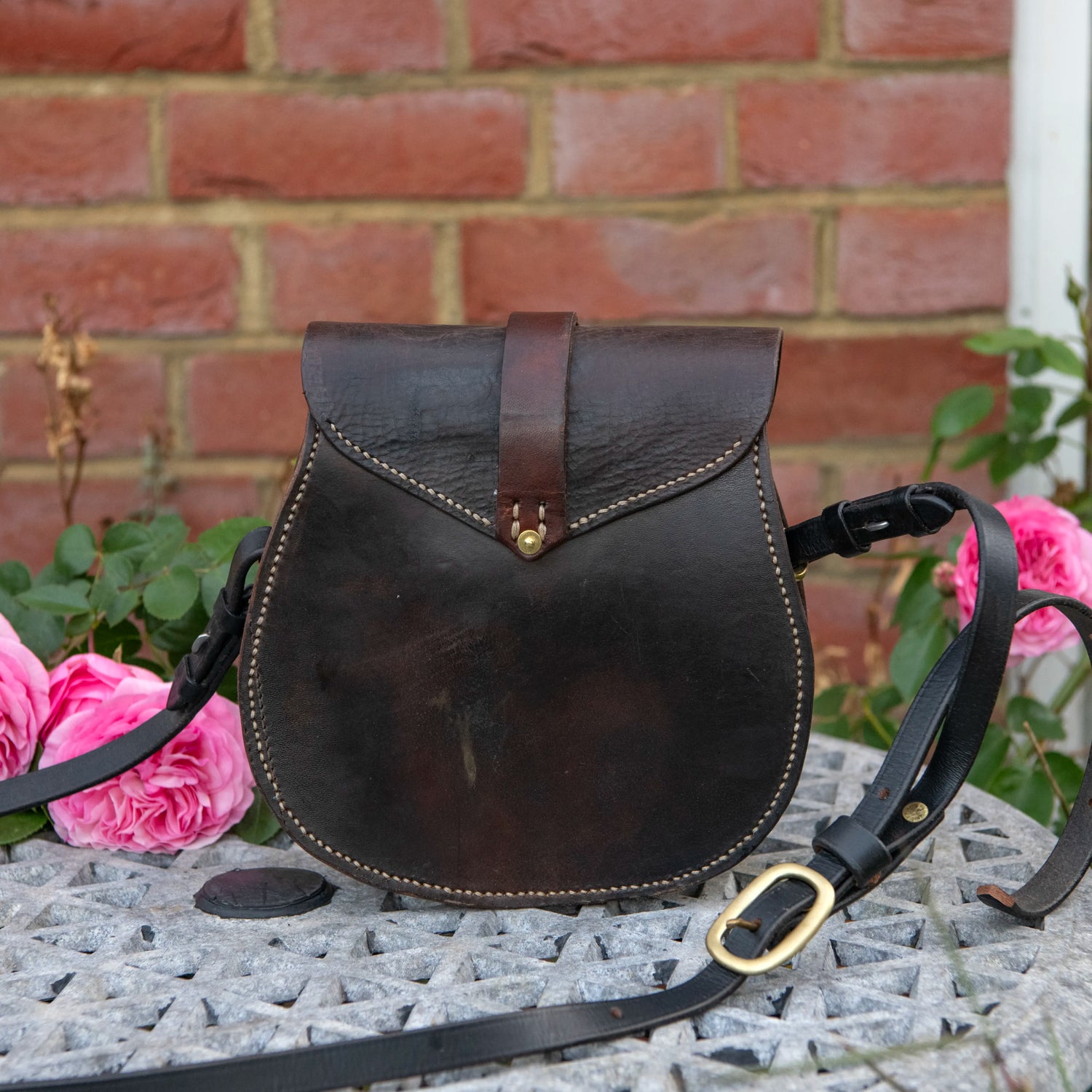 A straight-on environmental photograph showing the rear profile of The Uffington restored leather scrip resting upright on a stone table against a red brick wall, framed by pink roses. The image highlights the wide expanse and rich patina of the dark saddle leather, the V-shaped hand-stitching on the back panel, and the heavy equestrian shoulder strap sweeping across the foreground to display its solid brass adjustment buckle.