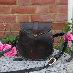 A straight-on environmental photograph showing the rear profile of The Uffington restored leather scrip resting upright on a stone table against a red brick wall, framed by pink roses. The image highlights the wide expanse and rich patina of the dark saddle leather, the V-shaped hand-stitching on the back panel, and the heavy equestrian shoulder strap sweeping across the foreground to display its solid brass adjustment buckle.