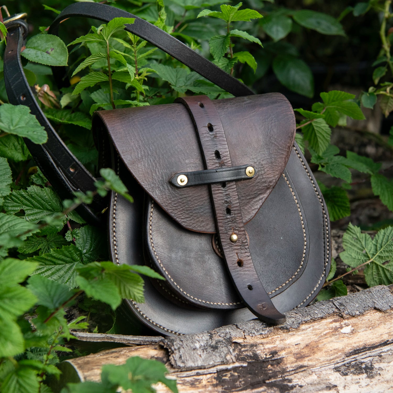 A dark-toned, moody environmental photograph of The Uffington leather scrip resting heavily upon a weathered log amidst natural green foliage. The image highlights the structured doublet construction, the central harness strap secured by a solid brass Sam Browne stud, and the rich, authentic grain of the restored vintage saddlery.