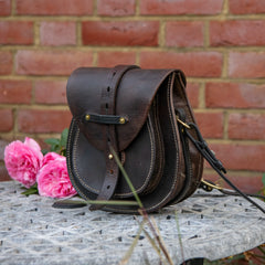 A three-quarter angle photograph of The Uffington restored leather scrip resting on a stone table against a red brick wall. Accompanied by pink roses, this side perspective reveals the structured depth and volume of the bag, clearly displaying the layered doublet construction, the side gusset, and the solid brass D-ring securing the heavy equestrian shoulder strap.