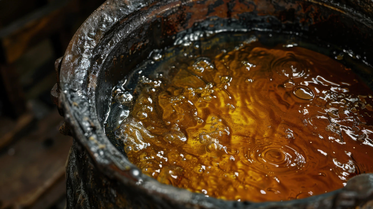 A moody, close-up photograph looking down into a heavily encrusted, rustic metal pot inside the Caracoda Equestrian workshop. A rich, natural mixture of beeswax and traditional conditioning oils is actively melting, bubbling, and swirling into a glowing, golden liquid. The atmospheric lighting highlights the viscous ripples on the surface, capturing the raw, authentic alchemy involved in hand-crafting our heritage leather restoration balms.