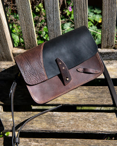 Front view of Oddity No. II, a unique two-tone repurposed saddle leather crossbody bag with a solid brass stud closure, resting in a natural outdoor setting.